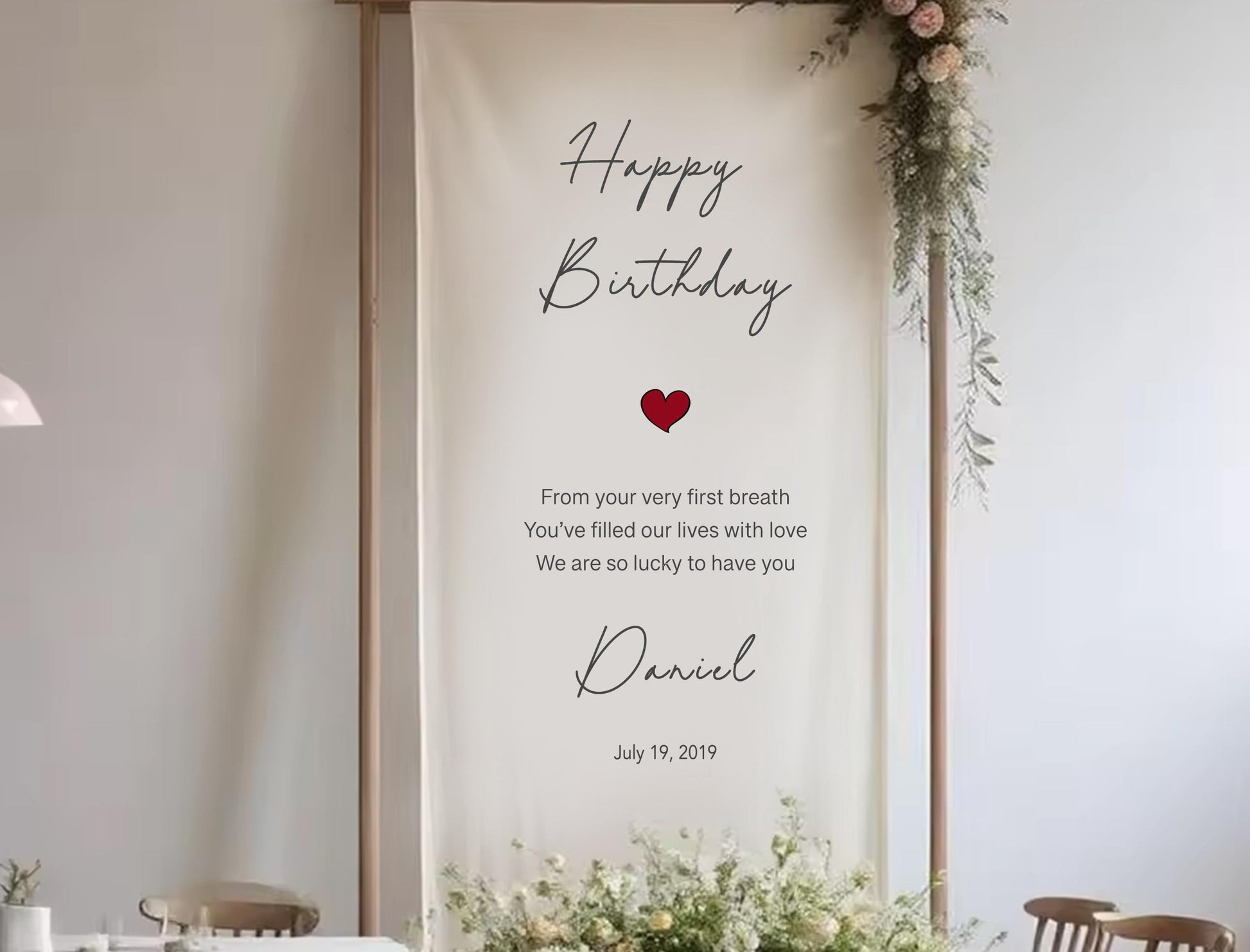 a banner with a birthday message for someone named Daniel. The banner has a white background with black text and a red heart symbol. The message reads: &quot;Happy Birthday Daniel. From your very first breath You&#39;ve filled our lives with love We are so lucky to have you&quot;. The banner is displayed in a room with a dining table and chairs, suggesting it is part of a celebration or event.