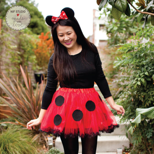 A woman wearing a Minnie Mouse-themed costume, including a red tutu with black polka dots, is standing in a garden surrounded by greenery.