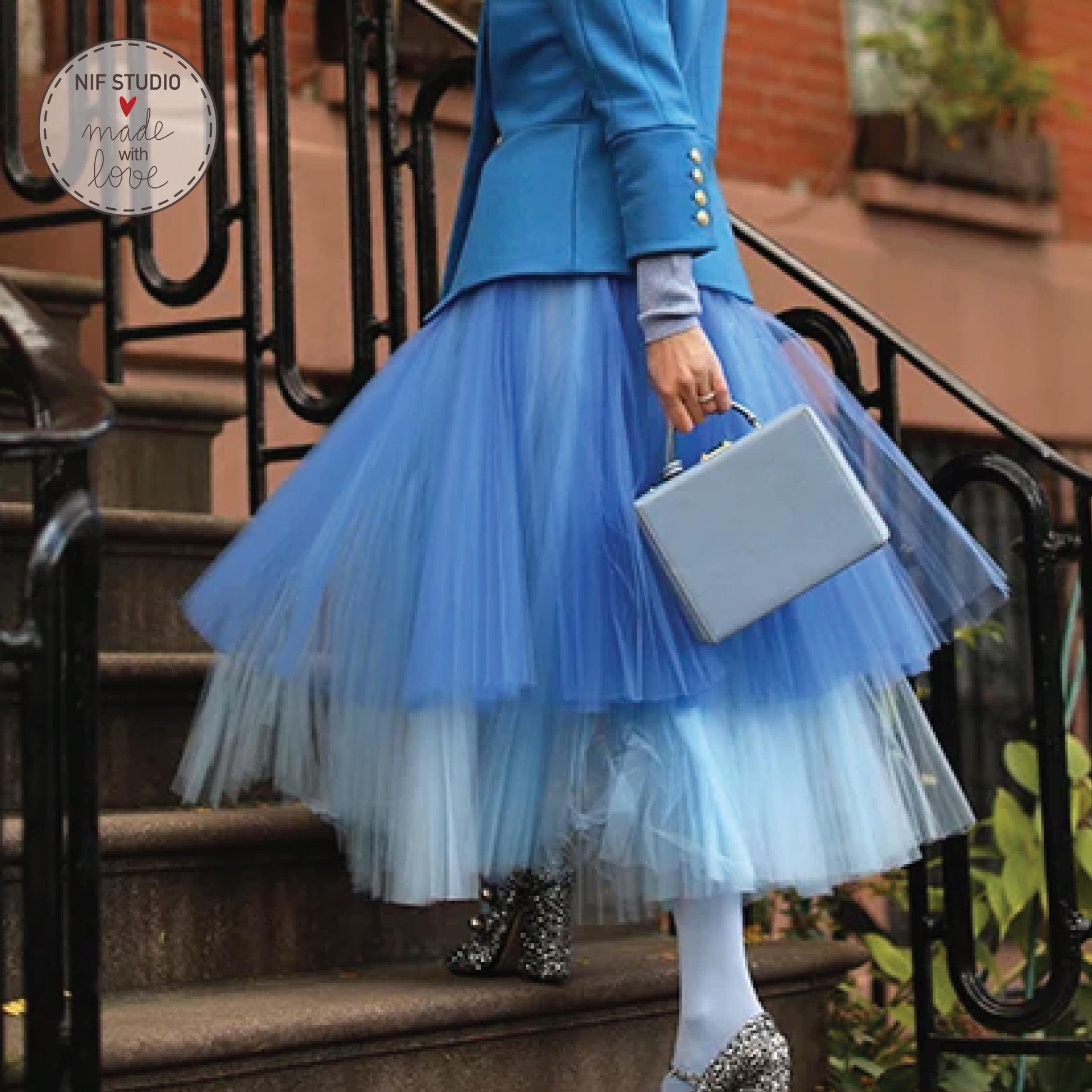 A woman wearing a blue jacket and a blue tulle skirt is walking down a staircase, carrying a small bag.