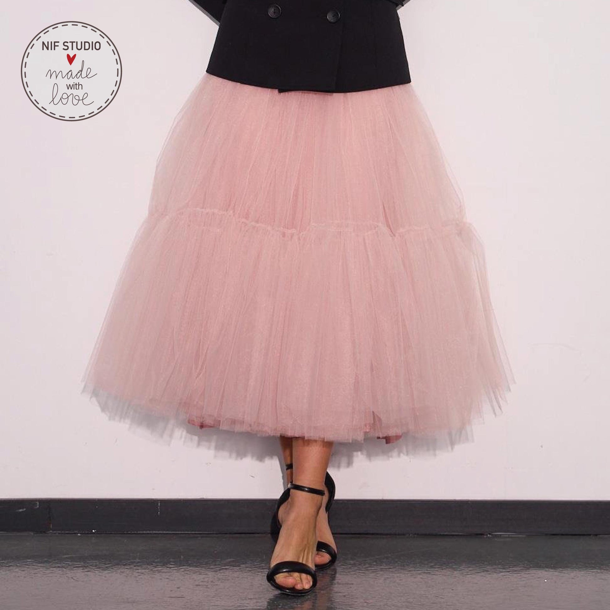 A woman wearing a pink tulle skirt and a black jacket stands against a white wall, with her feet visible in the foreground.