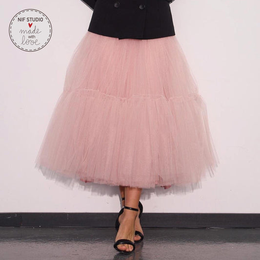 A woman wearing a pink tulle skirt and a black jacket stands against a white wall, with her feet visible in the foreground.