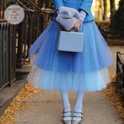 A woman wearing a blue tulle skirt and a blue jacket is holding a small gray purse and standing on a sidewalk.