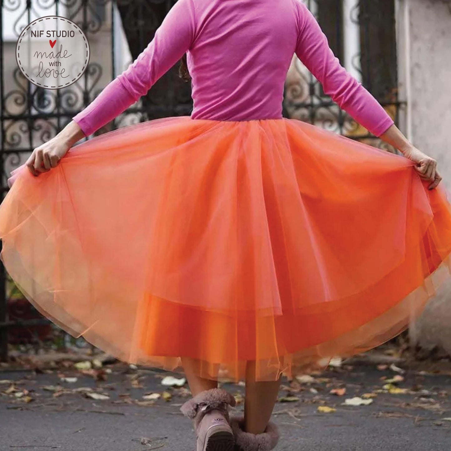 A woman wearing a pink top and an orange tulle skirt is standing in front of a wrought iron gate.