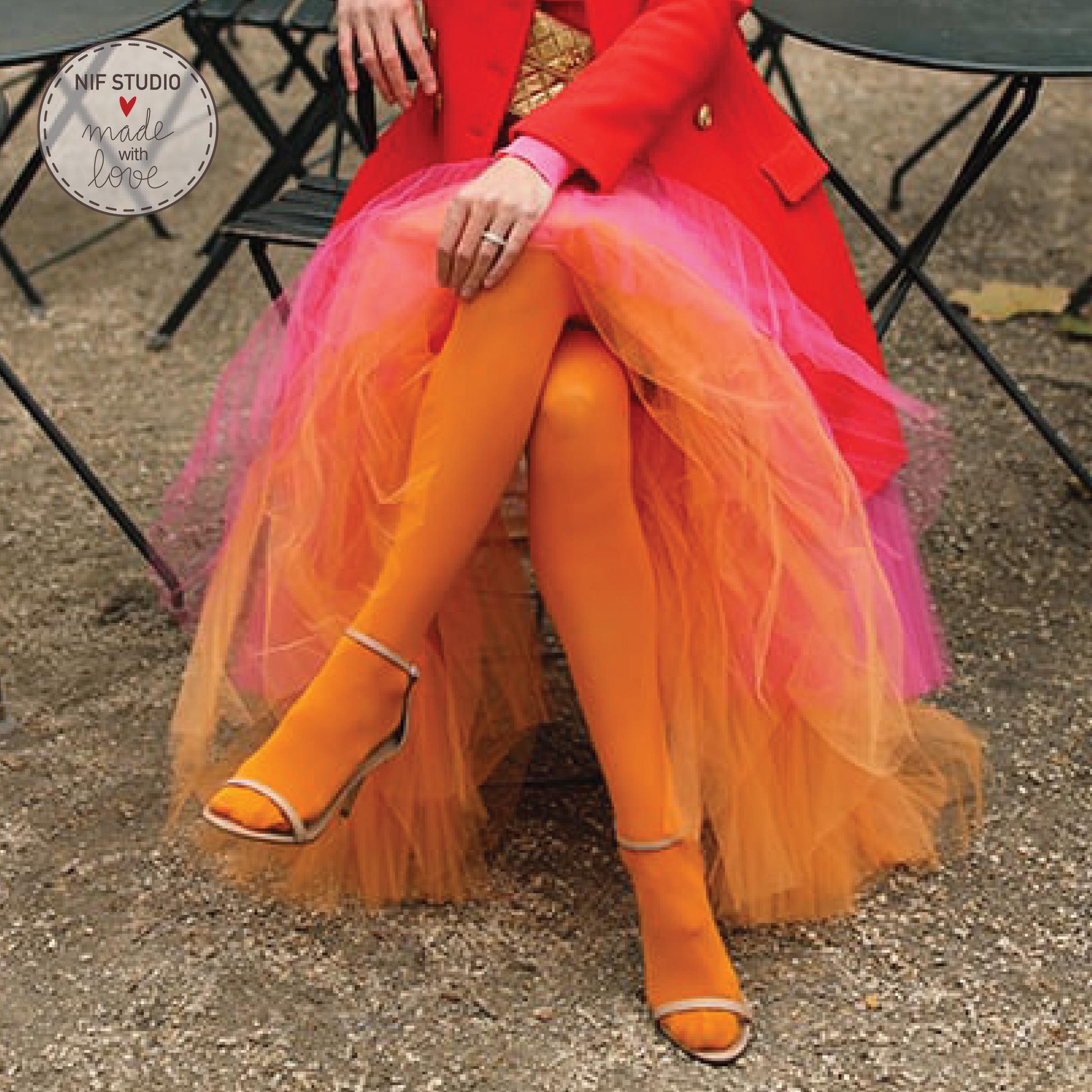 A woman wearing a red coat and a pink and orange tulle skirt is sitting on a chair, with her legs crossed.