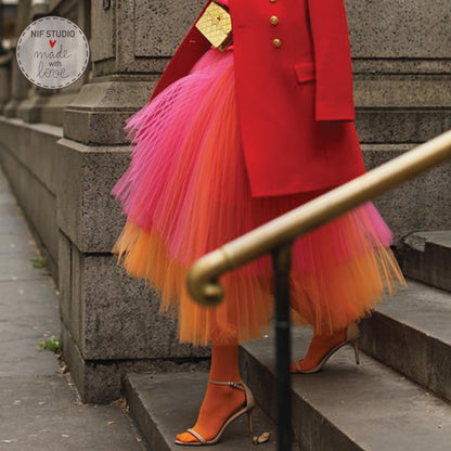A woman wearing a vibrant red coat and a colorful tulle skirt is walking down a set of stairs, with a pair of high heels visible.