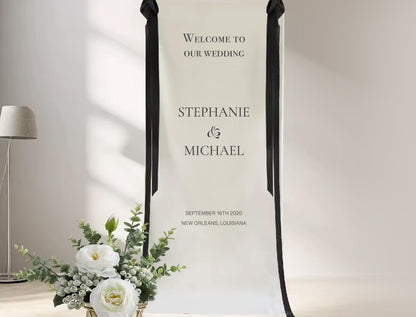 a white banner with black text welcoming guests to a wedding, along with the names &quot;Stephanie & Michael&quot; and the date &quot;September 16th, 2020&quot; in the background. There is also a vase of white flowers placed next to the banner.