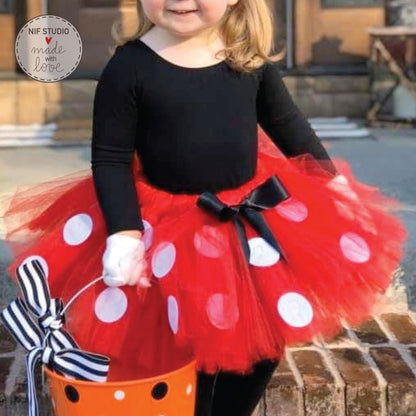 A young girl wearing a red and white polka dot tutu and holding a bucket with a striped ribbon, smiling at the camera.