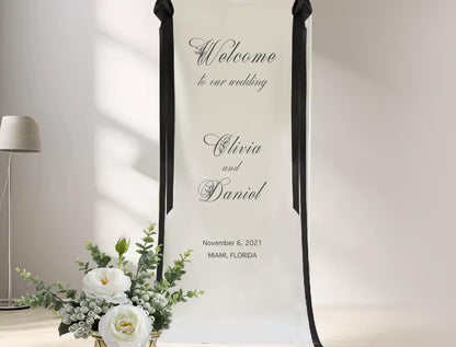 a white banner with black text that reads &quot;Welcome to our wedding&quot;, followed by the names &quot;Claire and Daniel&quot;. The banner also includes the date &quot;November 6, 2021&quot; and the location &quot;MAMMA, FLORIDA&quot;. In the foreground, there is a vase with white flowers and greenery, creating a pleasant and welcoming atmosphere for the wedding celebration.
